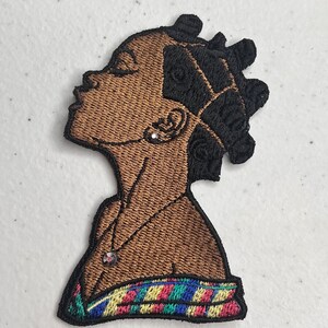 May include: Embroidered patch of a brown-skinned woman with black hair in a bun, wearing a colorful patterned scarf. The woman is looking up and has a necklace with a small, round pendant.