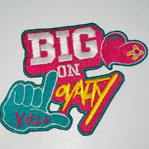 May include: Embroidered patch with the words "BIG ON LOYALTY" in white, yellow, and teal. The patch features a heart, a hand gesture, and the acronym "Y.O.L.O." on a pink background.
