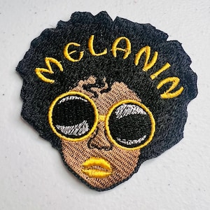 May include: Embroidered patch featuring a cartoon illustration of a person with an afro hairstyle wearing sunglasses. The person has brown skin and is wearing yellow sunglasses. The word "MELANIN" is embroidered in yellow lettering above the person's head.