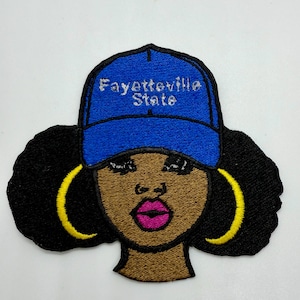 May include: Embroidered patch featuring a stylized Black woman wearing a blue baseball cap with the text "Fayetteville State". The woman has black hair, gold hoop earrings, and pink lipstick. The patch is on a white background.