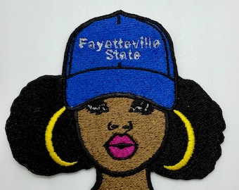 May include: Embroidered patch featuring a stylized Black woman wearing a blue baseball cap with the text "Fayetteville State". The woman has black hair, gold hoop earrings, and pink lipstick. The patch is on a white background.