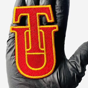 May include: Red felt letter "U" with a yellow outline. The letter is embroidered with a thick yellow thread.