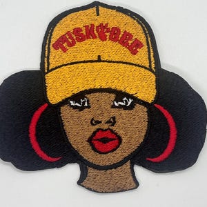 May include: Embroidered iron-on patch featuring a cartoon illustration of a Black woman with a yellow baseball cap that says "Tusky Babe" in red letters. She has large red hoop earrings and brown skin.