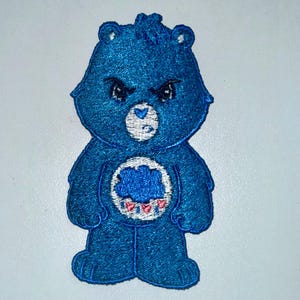 May include: A blue embroidered patch featuring a grumpy bear character. The bear has a dark blue body, a white belly emblem with a cloud and rain design, and a stern facial expression. The patch is likely for clothing or accessories.