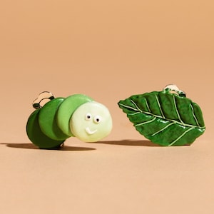May include: A pair of green and white caterpillar earrings and a green leaf earring. The caterpillar earrings have a smiling face.
