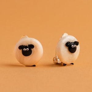 May include: A pair of white sheep-shaped earrings with black eyes and noses. The earrings are made of a smooth, white material and have a silver-toned metal backing.