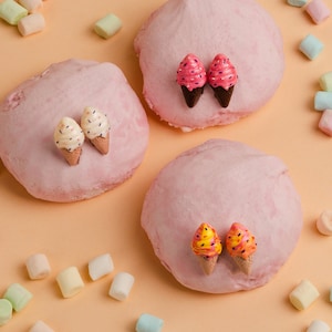 Puede incluir: Tres merengues rosas con pendientes de cono de helado de colores. Las merengues están rodeadas de malvaviscos de color pastel.