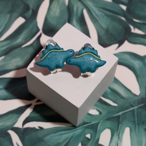 Cute turquoise little girls dinosaur - stegosaurus clip on earrings for unpierced ears in womens hands to show their size in real life, which is 2x1.4 cm or 0.79x0.55 inch.