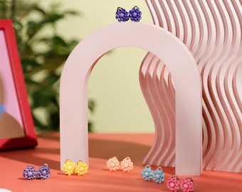 May include: A set of six pairs of flower-shaped earrings in various colors, including purple, yellow, pink, and blue. The earrings are displayed on a pink archway with a wavy background.