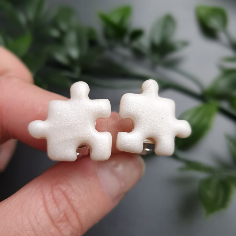 Marble white puzzle piece shape clip-on earring pair in women's hands to show size in real life which is 1.9cm or 0.75inch. Each piece has two protrusions and two recesses, and one of protrusions are pointing up and one to the side.