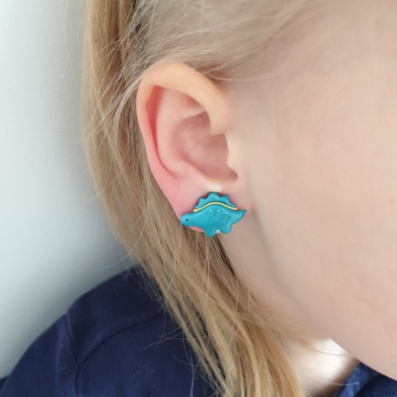 Turquoise stegosaurus, Dino earrings in 6 years old, blonde girls unpierced ears to show how they look in ear. Dinosaur is detailed with blue light line, and coupe of blue dots, black eye to make it more elaborate.