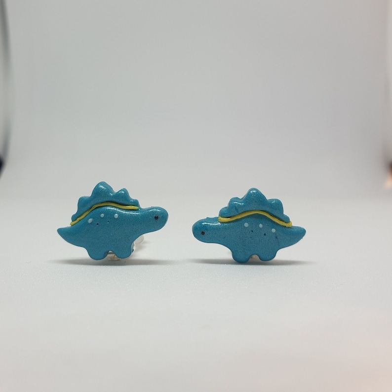 Cute turquoise little girls dinosaur - stegosaurus clip on earrings for unpierced ears in womens hands to show their size in real life, which is 2x1.4 cm or 0.79x0.55 inch.