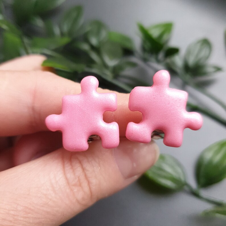 Light pink puzzle piece shape clip-on earring pair in women's hands to show size in real life which is 1.9cm or 0.75inch. Each piece has two protrusions and two recesses, and one of protrusions are pointing up and one to the side.