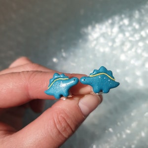 Cute turquoise little girls dinosaur - stegosaurus clip on earrings for unpierced ears in womens hands to show their size in real life, which is 2x1.4 cm or 0.79x0.55 inch.