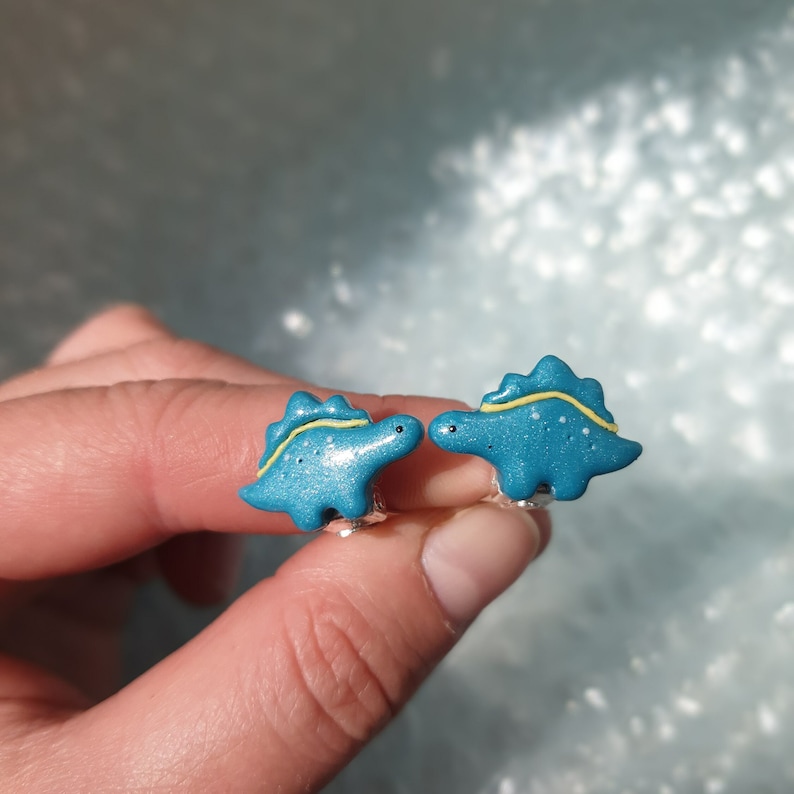 Cute turquoise little girls dinosaur - stegosaurus clip on earrings for unpierced ears in womens hands to show their size in real life, which is 2x1.4 cm or 0.79x0.55 inch.