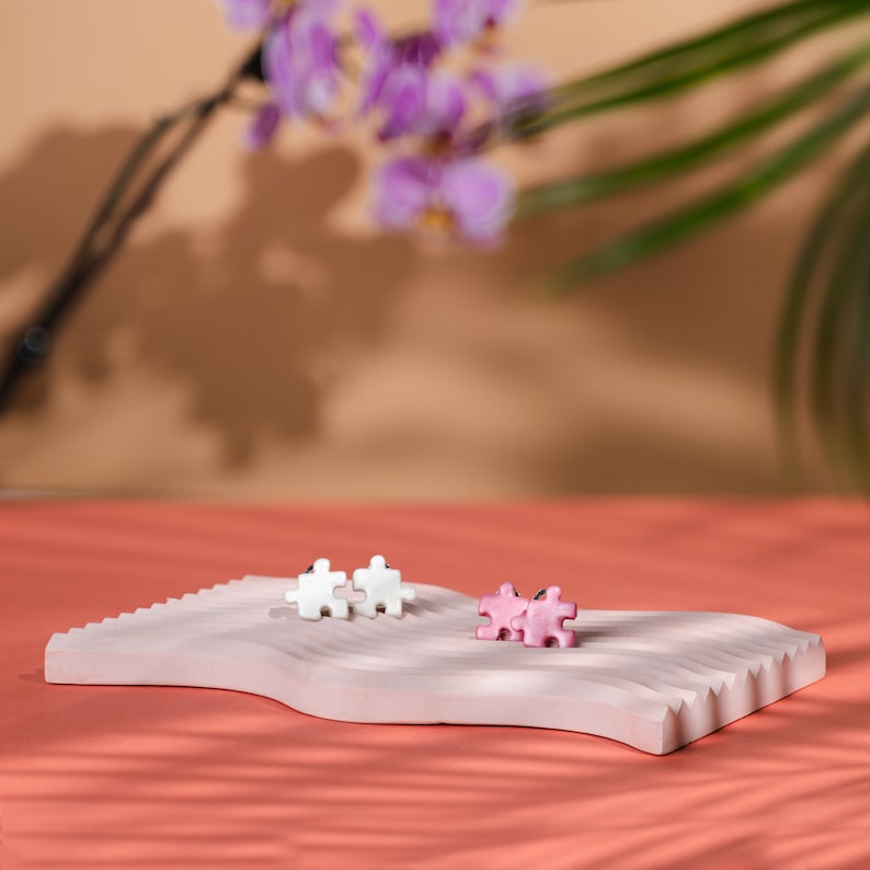 Pink and white puzzle piece shape clip-on earring pairs. Each puzzle piece has two protrusions and two recesses, and one of protrusions are pointing up and one to the side. White pair is marble white color. Pink is light pink color with little shine