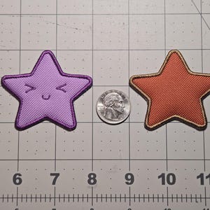 May include: Two star-shaped fabric patches. One is purple with a smiling face and a dark purple border. The other is orange with a gold border. A quarter coin is between the patches. The patches are on a grid.