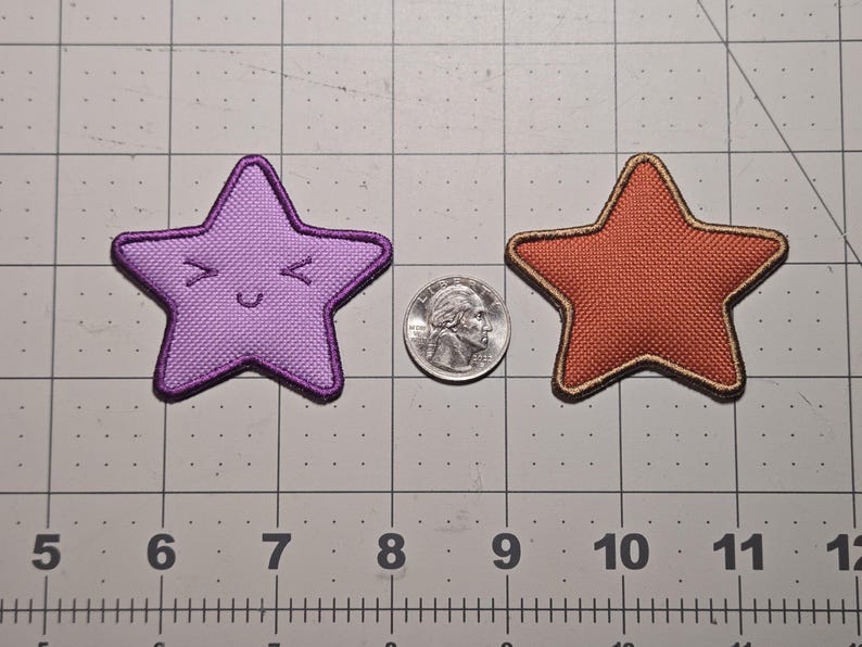 May include: Two star-shaped fabric patches. One is purple with a smiling face and a dark purple border. The other is orange with a gold border. A quarter coin is between the patches. The patches are on a grid.