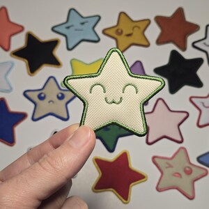 May include: A hand holding a beige star-shaped patch with a green outline and a smiling face. Other star patches in various colors and expressions are scattered in the background, including blue, red, and black.