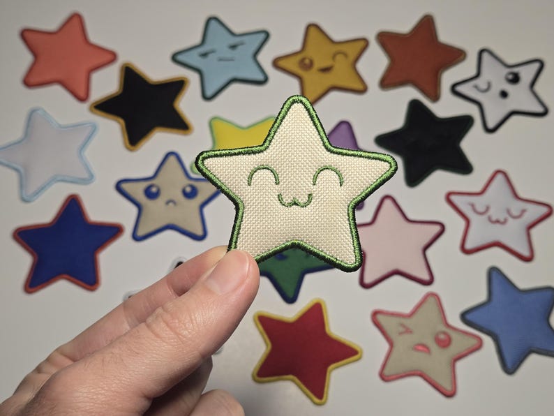 May include: A hand holding a beige star-shaped patch with a green outline and a smiling face. Other star patches in various colors and expressions are scattered in the background, including blue, red, and black.