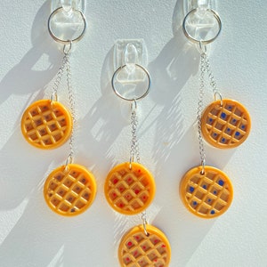 May include: Three keychains with waffle-shaped charms. Each charm is a different colour: yellow with red dots, yellow with blue dots, and yellow with brown dots.