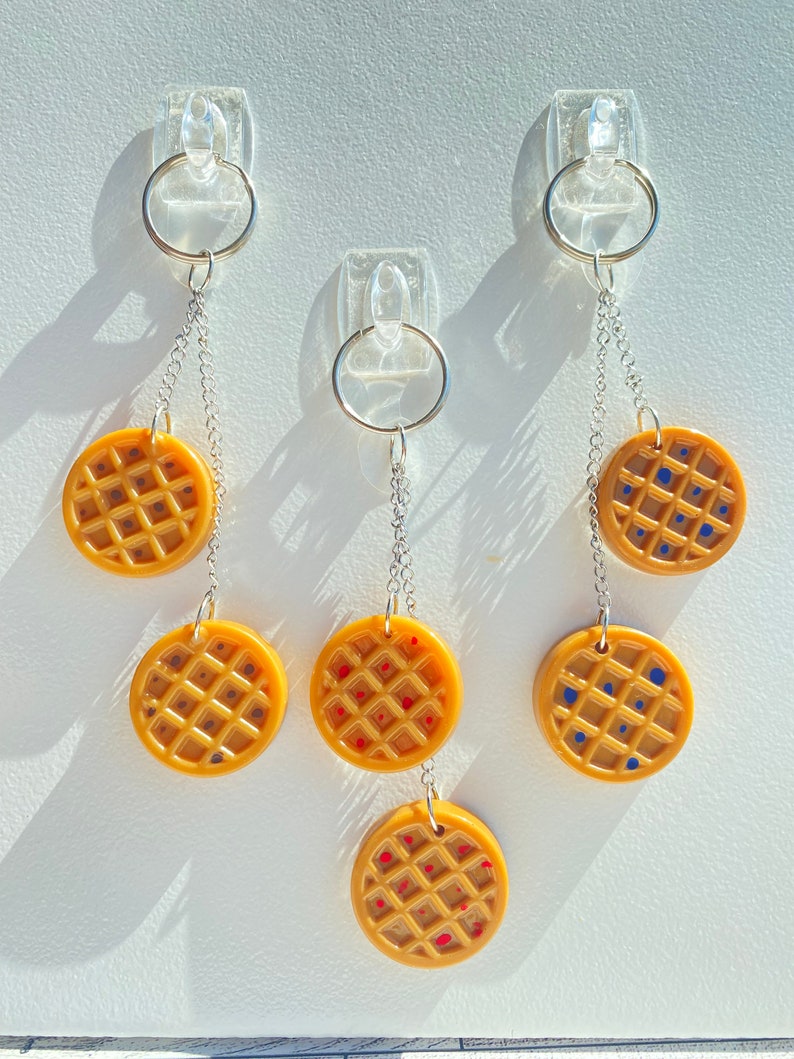 May include: Three keychains with waffle-shaped charms. Each charm is a different colour: yellow with red dots, yellow with blue dots, and yellow with brown dots.