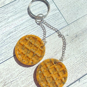 May include: A keychain with two round, yellow waffle charms featuring a black poppy seed design. The charms are attached to a silver chain and a silver key ring.