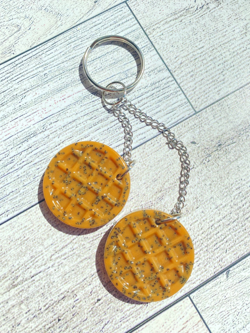 May include: A keychain with two round, yellow waffle charms featuring a black poppy seed design. The charms are attached to a silver chain and a silver key ring.