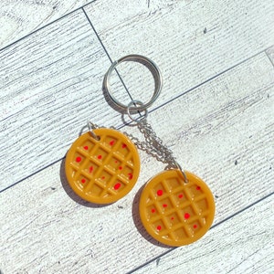 May include: A keychain featuring two round, brown waffle-shaped charms with red syrup-like accents. The charms are attached to a silver chain and a silver key ring.