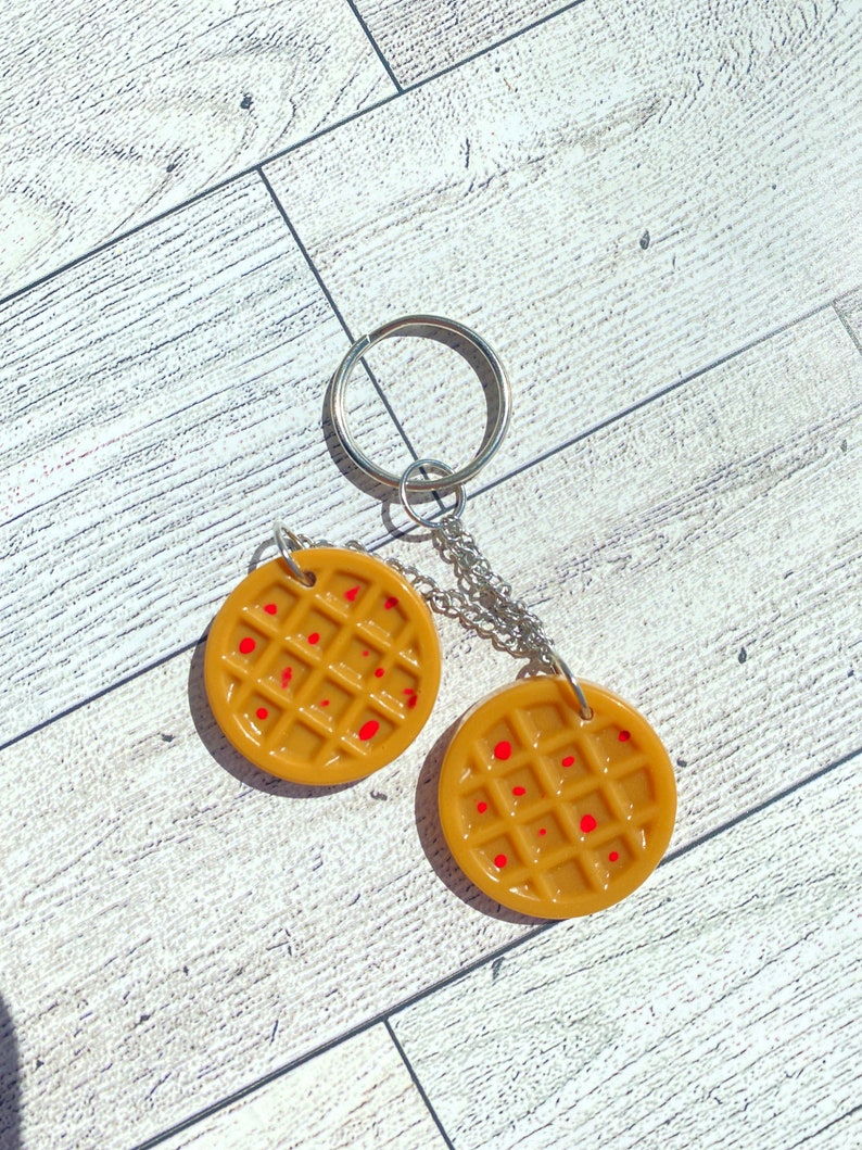 May include: A keychain featuring two round, brown waffle-shaped charms with red syrup-like accents. The charms are attached to a silver chain and a silver key ring.