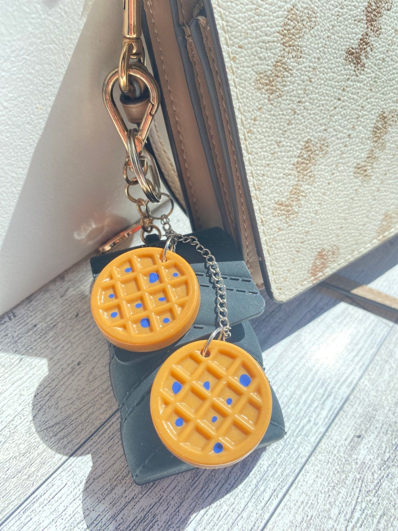 May include: A gold chain keychain with two round, brown waffle-shaped charms. The charms have blue dots on them.
