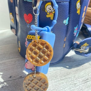 May include: A blue and white hand sanitiser holder with a keychain attached. The keychain has two brown waffle-shaped charms and a silver chain. The holder has the text "COOKY @BT21" printed on it.