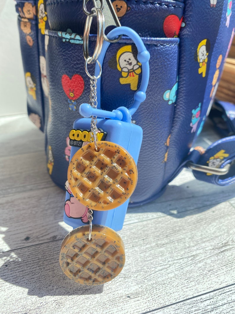 May include: A blue and white hand sanitiser holder with a keychain attached. The keychain has two brown waffle-shaped charms and a silver chain. The holder has the text "COOKY @BT21" printed on it.