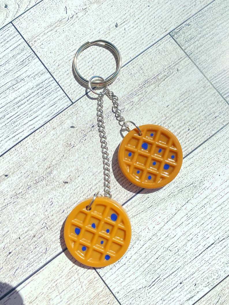 May include: A keychain with two waffle-shaped charms. The charms are brown with blue spots and are attached to a silver chain and ring.