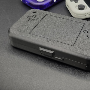 May include: Three handheld gaming consoles. One is black with a textured surface, one is white with a black screen, and one is purple with a black screen.