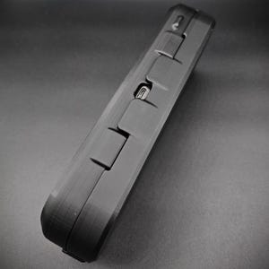 May include: A black, rectangular case with a textured surface. The case features several raised rectangular sections and a small port. The case is angled, showing its side and top.