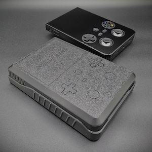 以下が含まれることがあります： 2つの黒い携帯ゲーム機が表示されています。下のコンソールは、エンボス加工されたボタンと「ANBERNIC」の文字が付いたテクスチャ加工された表面を持っています。上のコンソールは、コントローラーレイアウトで滑らかです。コンソールは濃い灰色の表面にあります。