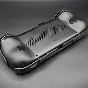 May include: Black handheld gaming device case with textured grips and button details. The case has a perforated vent area and a sleek, ergonomic design. The device is designed to enhance the gaming experience.