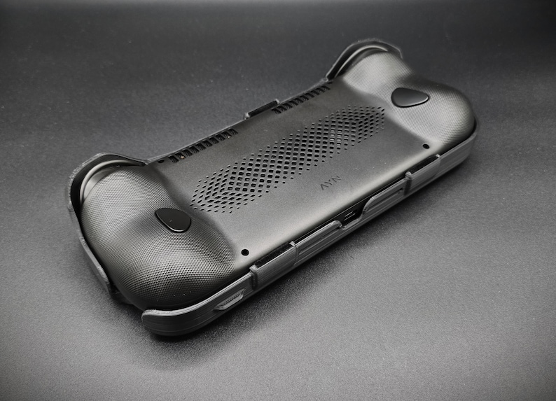 May include: Black handheld gaming device case with textured grips and button details. The case has a perforated vent area and a sleek, ergonomic design. The device is designed to enhance the gaming experience.