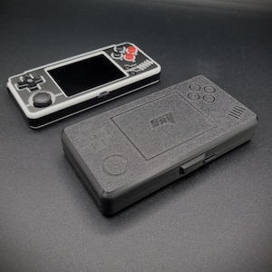 May include: Two handheld video game consoles, one black and white with red buttons and a screen, the other black with a textured surface and a circular button.