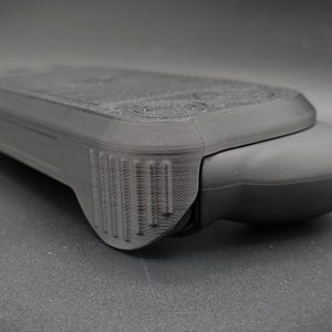 May include: A dark gray, rectangular electronic device with a rounded end. The device has a textured surface and a series of vertical lines on one side. The overall design suggests a handheld gadget or controller.