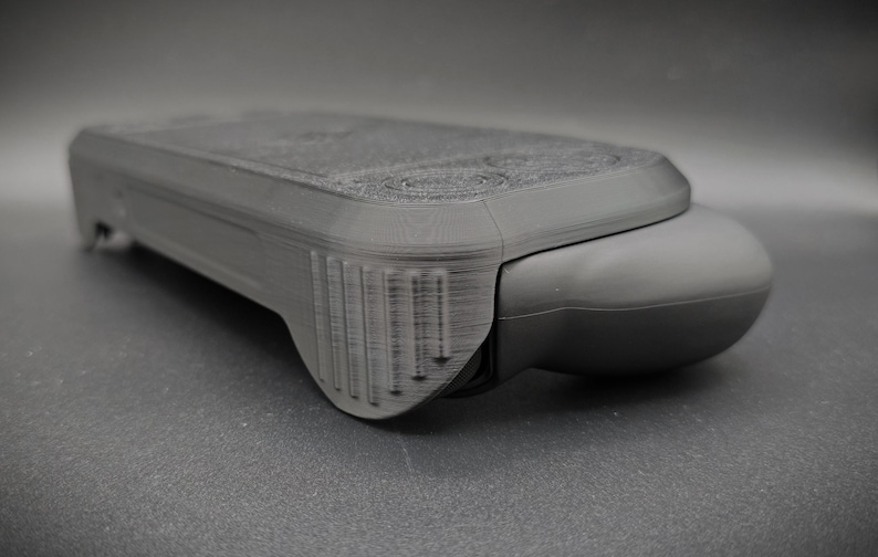 May include: A dark gray, rectangular electronic device with a rounded end. The device has a textured surface and a series of vertical lines on one side. The overall design suggests a handheld gadget or controller.