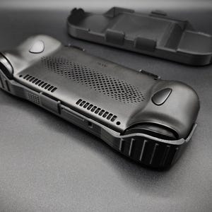 May include: A black, textured handheld gaming device case with a matching detached top piece. The case features a series of vents, buttons, and a textured grip. The design is sleek and modern, with a focus on functionality and protection.