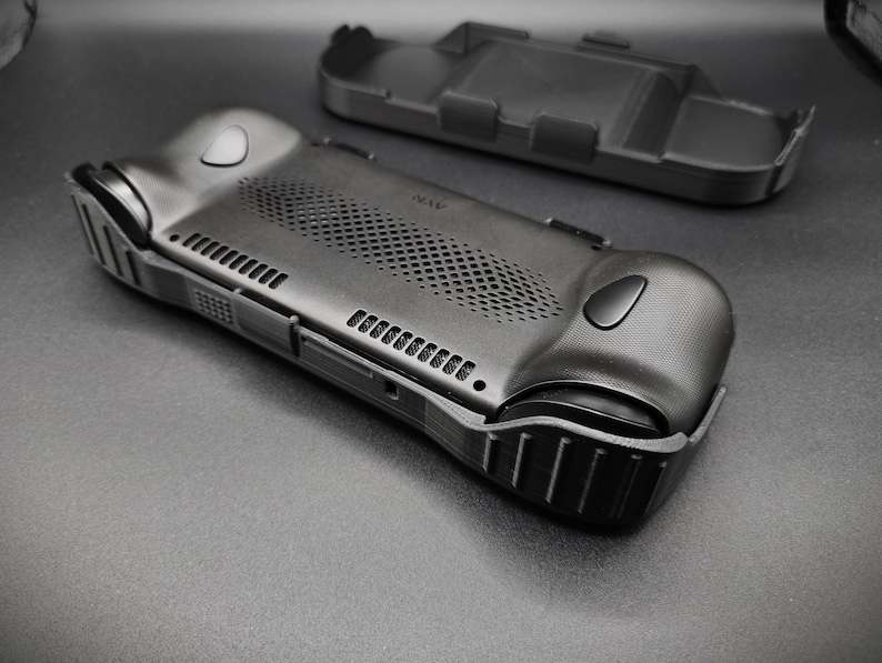 May include: A black, textured handheld gaming device case with a matching detached top piece. The case features a series of vents, buttons, and a textured grip. The design is sleek and modern, with a focus on functionality and protection.