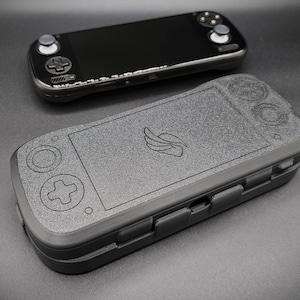 Könnte beinhalten: Zwei schwarze Handheld-Gaming-Geräte werden gezeigt. Eines ist eine schlanke Konsole mit Bildschirm und Tasten. Das andere ist ein strukturiertes Gehäuse mit Controller-Layout und Logo. Das Gehäuse scheint zum Schutz der Konsole bestimmt zu sein.