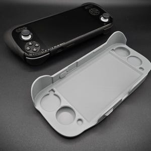 May include: A black handheld gaming console with a gray protective case. The console features a screen, buttons, and control sticks. The gray case is designed to fit the console, with openings for the buttons and screen.