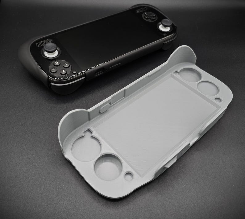 May include: A black handheld gaming console with a gray protective case. The console features a screen, buttons, and control sticks. The gray case is designed to fit the console, with openings for the buttons and screen.