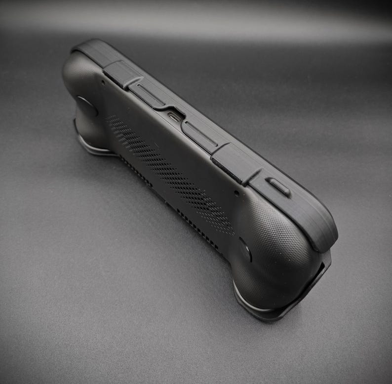 May include: A black handheld gaming device with a textured surface and multiple buttons. The device has a curved design with ergonomic grips on each side. The top edge features additional buttons and a USB-C port.