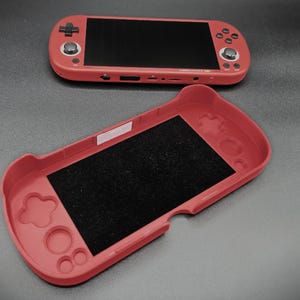 以下が含まれることがあります： 2つの赤い携帯ゲーム機が表示されています。1つは開いており、黒い内部が見え、もう1つは黒い画面で閉じられています。コンソールにはボタンと方向パッドがあります。