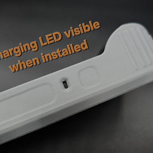 May include: A close-up of a white, rectangular device with a ridged top and a small, dark port. The text "Charging LED visible when installed" is in orange. The device appears to be made of plastic.